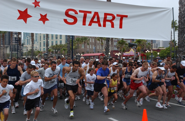 Eine Gruppe von Marathonläufern läuft an einer Verkehrsinsel mit einem Banner und Bäumen im Hintergrund vorbei unter einem klaren blauen Himmel.