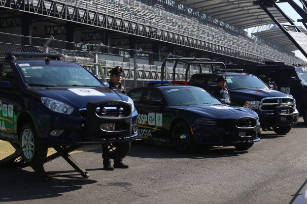 Eine Gruppe von Polizeiwagen steht vor einer Rennstrecke, uniformierte Personen stehen auf der Straße, im Hintergrund ist ein Stadion und ein Maschendrahtzaun zu sehen.