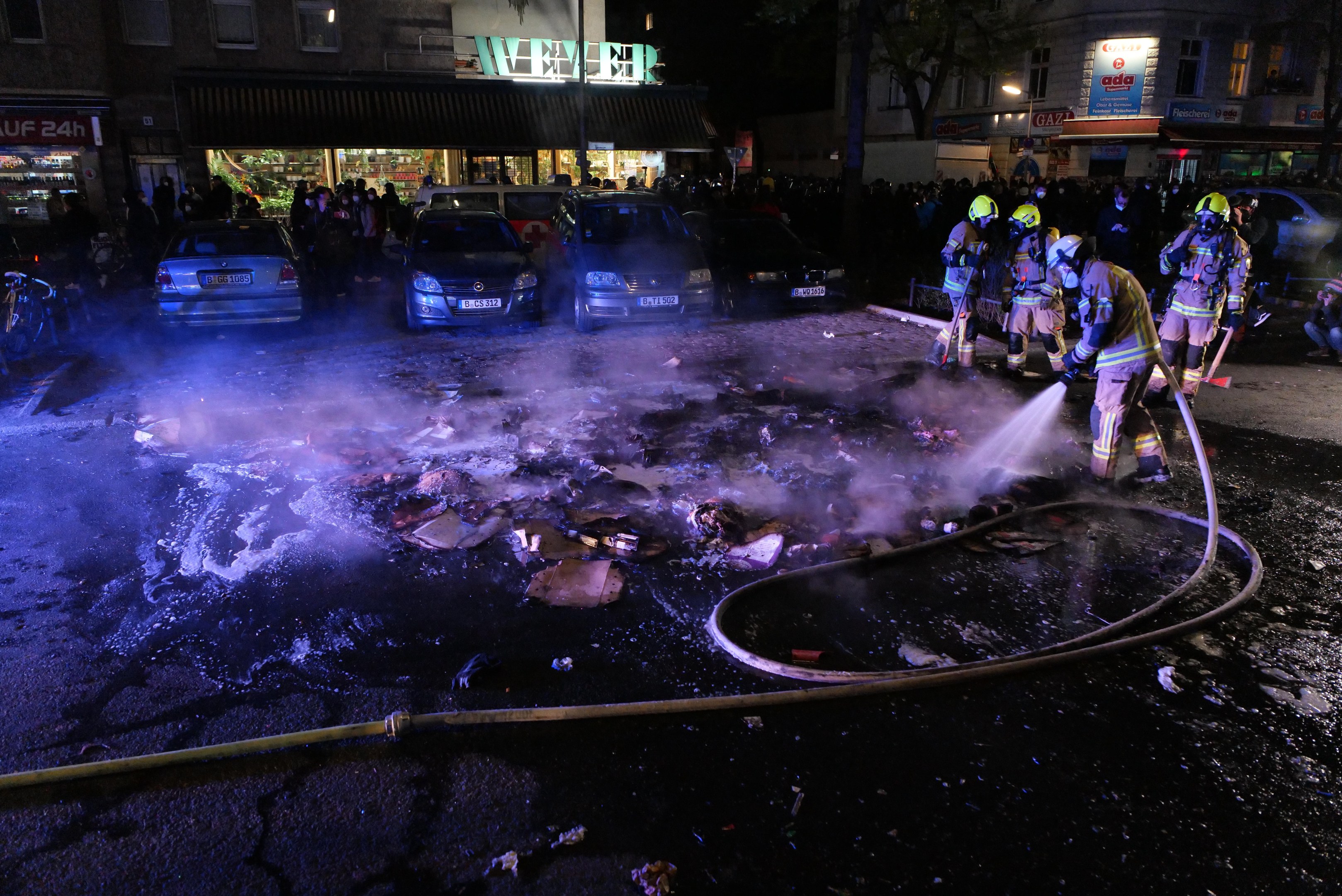 Feuerwehrleute in Helmen stehen um ein Straßenfeuer herum, halten Rohre, mit Fahrzeugen, einem Rohr auf dem Boden, Gebäuden, Namensschildern, Laternenpfählen, Bäumen und einem dunklen Himmel im Hintergrund.