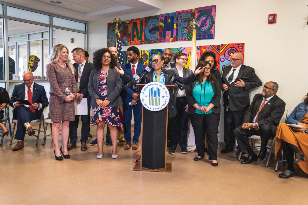 Eine Gruppe von Menschen steht vor einem Podium mit einem Mikrofon und einer Tafel mit Text, während andere auf Stühlen sitzen oder in der Nähe stehen; Flaggen, ein Feuerlöscher und Deckenlampen sind an der Rückwand zu sehen.
