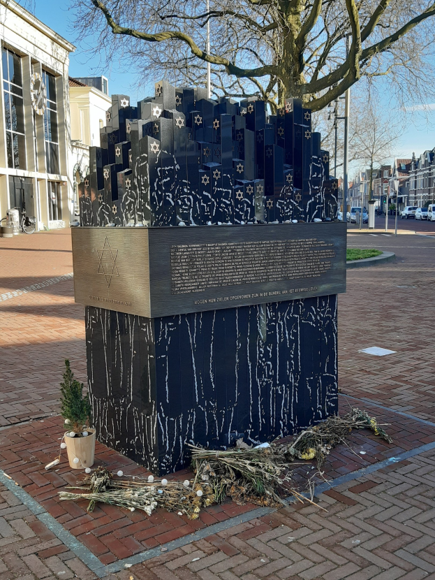 Denkmal für Holocaust-Opfer in einem Stadtplatz, mit einem Baum, Topfpflanze, verstreuten Blättern und urbaner Hintergrund.