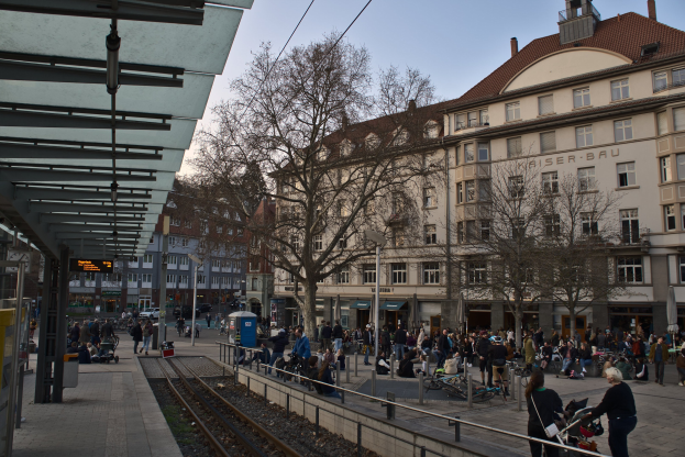 Eine belebte Stadtstraße mit einem Bahnhof auf der linken Seite, Menschen, die gehen, auf Bänken sitzen und stehen, umgeben von Gebäuden, Bäumen, Laternenmasten und Texttafeln unter einem sichtbaren Himmel.