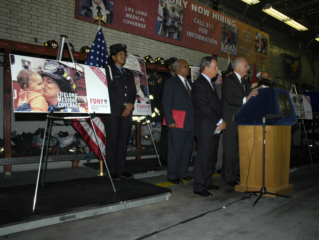 Eine Gruppe von Menschen steht in der N├Ąhe eines Podiums mit Mikrofonen, Schildern auf St├Ąnden, Fahnen und Schildern an der Wand, alles unter hellen Lichtern.