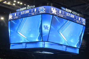 Ein großer Bildschirm in der Mitte eines Basketballfeldes zeigt "Kentucky Wildcats vs Florida State Seminoles", umgeben von einer Gruppe von Menschen auf den Tribünen und einer Decke mit Lampen darüber.