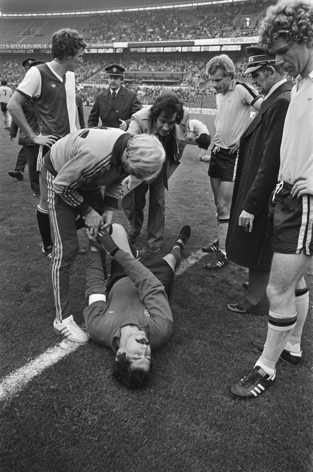 Schwarzes und weißes Bild eines Fußballspielers, der auf dem Boden liegt und von Mannschaftskameraden und einem Stadionpublikum umgeben ist, der scheinbar verletzt ist.