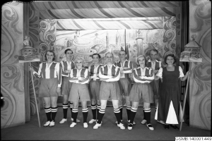 Schwarzes und weißes Foto einer Frauenfußballmannschaft der 1940er Jahre, die auf einer Bühne stehen, lächeln und Stöcke halten, mit einer bemalten Wand und Text unten.