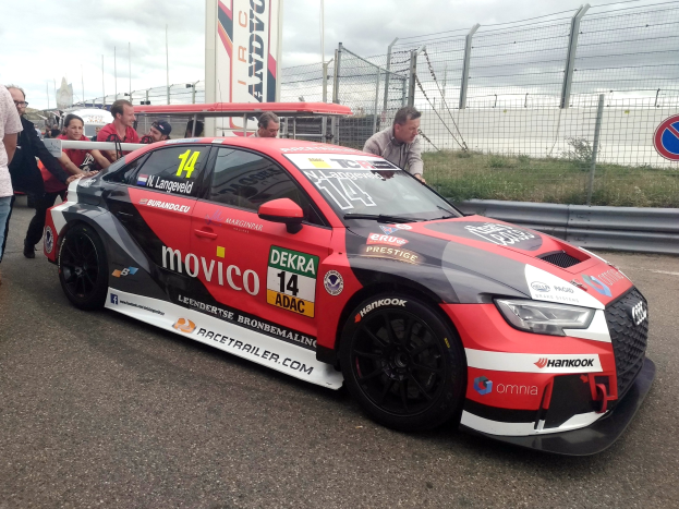 Ein Audi RS3 DTM-Rennwagen auf der Straße, umgeben von einer Gruppe von Menschen, mit einem Zaun, Gras, einem Schild, einer Fahne mit Text, Pfosten und einem bewölkten Himmel im Hintergrund.