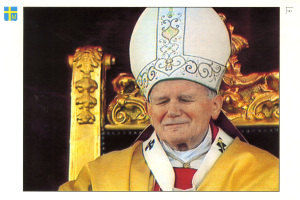 Ein Mann in priesterlicher Robe und weißer Mütze sitzt auf einem Stuhl vor dunklem Hintergrund und strahlt Würde und Ernsthaftigkeit aus.