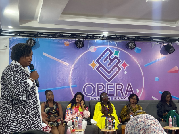 Frau vor sitzenden Frauen auf einer Couch stehend, Mikrofon haltend, mit Flaschen und Gegenständen auf Tischen im Vordergrund, Banner mit Text und Deckenbeleuchtung im Hintergrund.