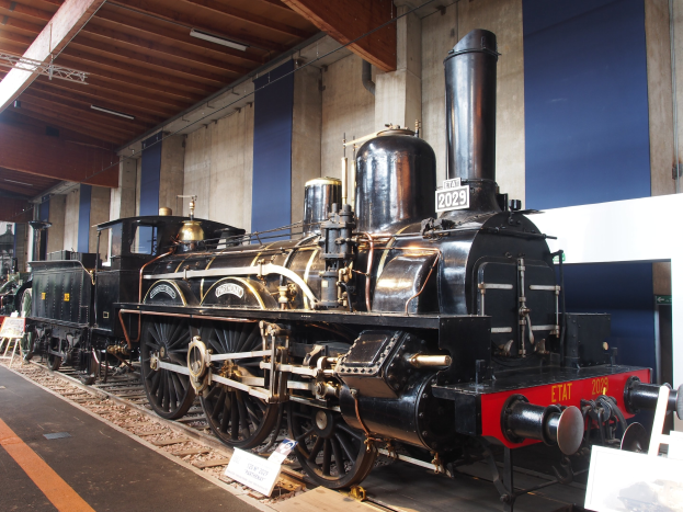 Alte Dampflokomotive in einem Museum ausgestellt mit Eisenbahnschienen, Informationsschildern und anderen Ausstellungsstücken gegen eine Wand mit Deckenleuchten.