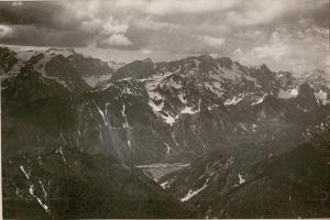 Ein Schwarz-Weiß-Foto einer Gebirgskette mit schneebedeckten Gipfeln und einem bewölkten Himmel, mit dem Text "Aussicht auf die Alpen vom Gipfel des Berges" unten.