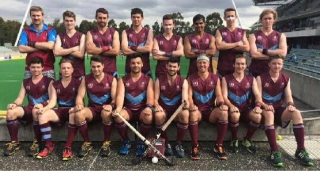 Eine Gruppe von Männern in Sportuniformen steht zusammen auf einem Feld, einige halten Hockey-Schläger und feiern mit erhobenen Armen vor einem Stadion mit Zuschauern und einer Kulisse aus Bäumen unter einem klaren blauen Himmel.