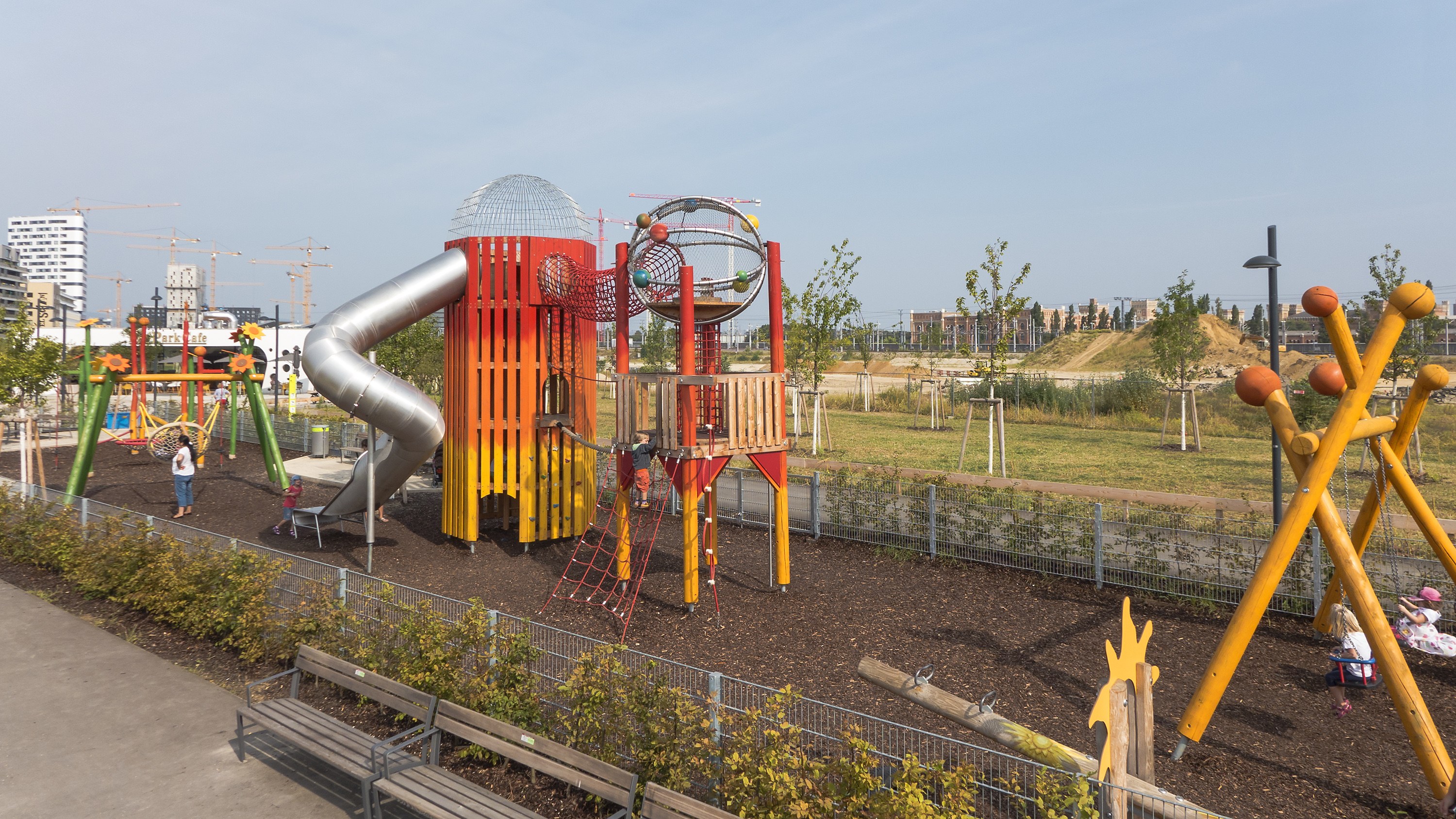 Ein Spielplatz mit buntem Equipment, einschließlich einer Rutsche und Schaukeln, umgeben von Pflanzen, einem Zaun, Bänken und einer Straße, mit Bäumen, Gebäuden und Kränen im Hintergrund unter einem sichtbaren Himmel.