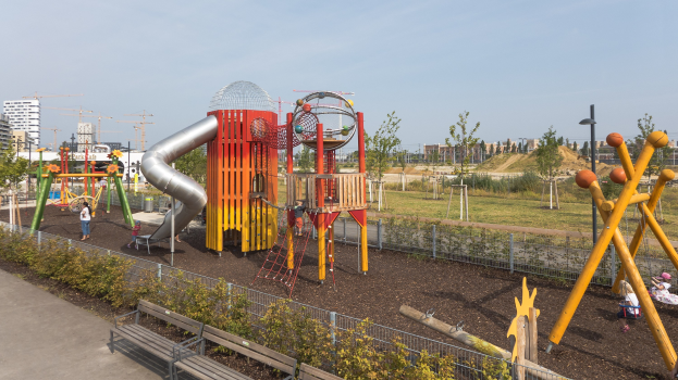 Ein Spielplatz mit buntem Equipment, einschließlich einer Rutsche und Schaukeln, umgeben von Pflanzen, einem Zaun, Bänken und einer Straße, mit Bäumen, Gebäuden und Kränen im Hintergrund unter einem sichtbaren Himmel.