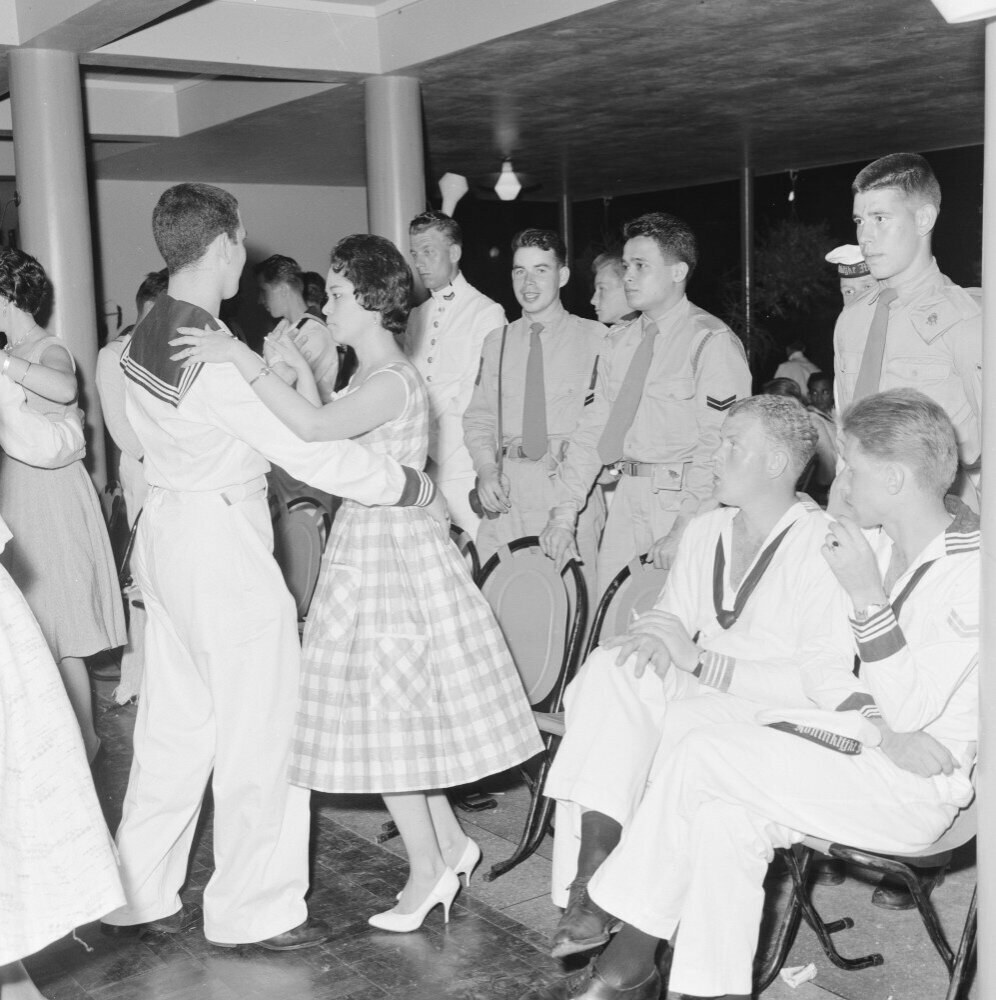 Schwarz-weißes Bild von Militärpersonal, das auf einer Tanzfläche tanzt, einige sitzen und einige stehen, mit Säulen und Lichtern im Hintergrund.
