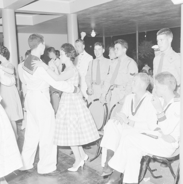 Schwarz-weißes Bild von Militärpersonal, das auf einer Tanzfläche tanzt, einige sitzen und einige stehen, mit Säulen und Lichtern im Hintergrund.