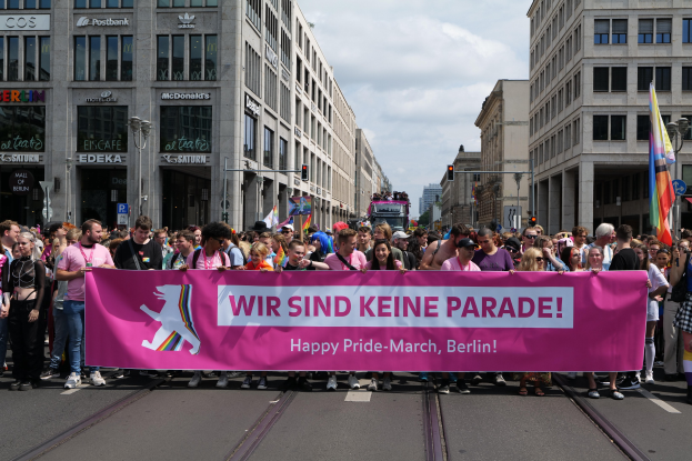 Eine Gruppe von Menschen marschiert auf einer Berliner Straße und hält ein pinkfarbenes Banner mit der Aufschrift "Happy Pride March", während Gebäude, Laternenpfähle und Verkehrszeichen die Straße säumen.