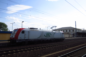 Ein Diesel-Elektrolokomotive fährt auf Schienen neben einem Gebäude, mit Strommasten, Kabeln, Straßenlaternen, Bäumen und einem bewölkten Himmel im Hintergrund.