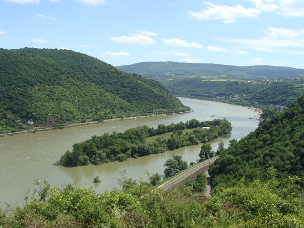 Panoramablick auf den Rhein, der sich durch eine grüne Landschaft schlängelt, mit einer Straße daneben, unter einem teilweise bewölkten Himmel.