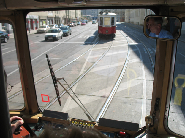 Mann fährt Straßenbahn auf einer Stadtstraße mit anderen Fahrzeugen und Gebäuden im Hintergrund bei klarem blauem Himmel.