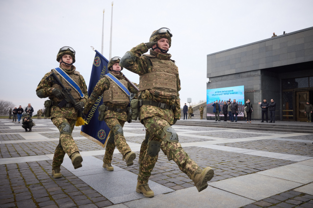 Gruppe ukrainischer Soldaten marschiert in Formation auf einer Straße, trägt militärische Uniformen und trägt Gewehre und Flaggen, mit Zivilisten und städtischen Elementen im Hintergrund.