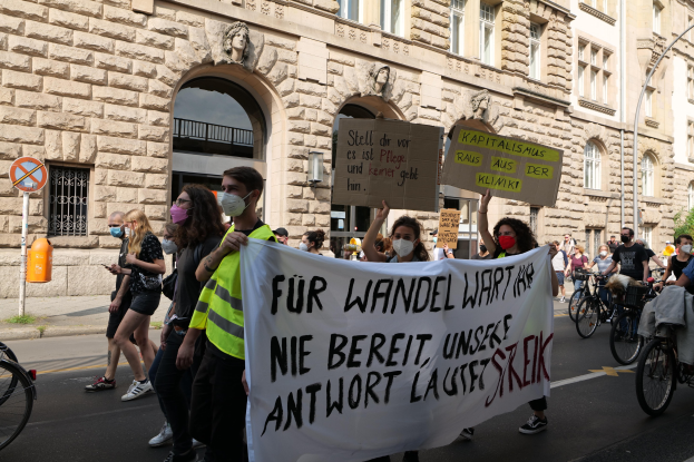 Eine Gruppe von Menschen marschiert auf einer Demonstration gegen die Anti-Kriegs-Bewegung in Berlin, einige halten Schilder und andere fahren Fahrräder, im Hintergrund ist ein Gebäude mit Fenstern, Bögen, Säulen und Skulpturen zu sehen.