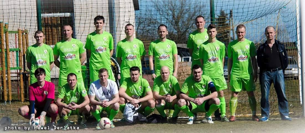 Eine Gruppe von Männern in grünen T-Shirts steht auf einem Fußballfeld mit einem Ball davor, einige sind in der Hocke, in der Nähe eines Torpfostens mit Bäumen und Gebäuden im Hintergrund.