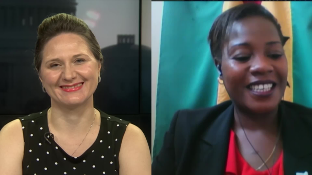 Zwei Frauen, die während eines Videoanrufs lächeln, eine in einem schwarzen und weißen Kleid, die andere in einer unbekannten Farbe, mit einer Flagge an der Wand hinter ihnen an einem öffentlichen Ort.