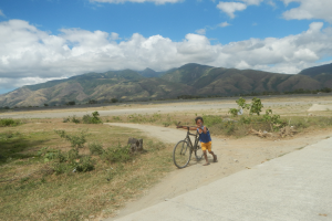 Ein junger Junge fährt ein Fahrrad auf einem Schotterweg durch eine grüne Landschaft mit Bergen unter einem blauen Himmel mit weißen Wolken.