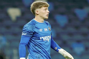 Ein junger Mann in einem blauen Torwarttrikot, einschließlich eines blauen T-Shirts und weißer Handschuhe, steht auf einem Fußballfeld während eines Spiels, mit einem unscharfen Hintergrund.