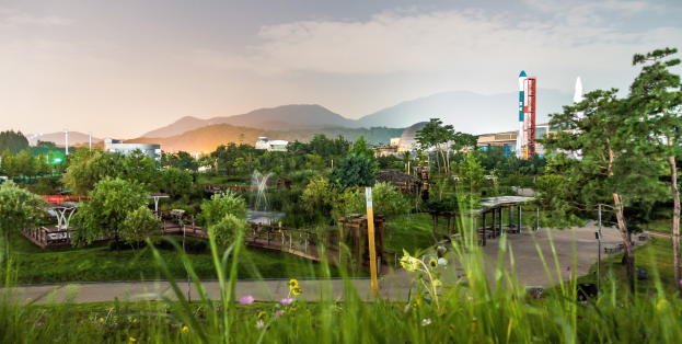 Ein Park mit grünem Gras, bunten Blumen, hohen Bäumen, einer Brücke mit Geländern, einem Springbrunnen, Laternenmasten, Gebäuden im Hintergrund, majestätischen Bergen und einem klaren blauen Himmel.