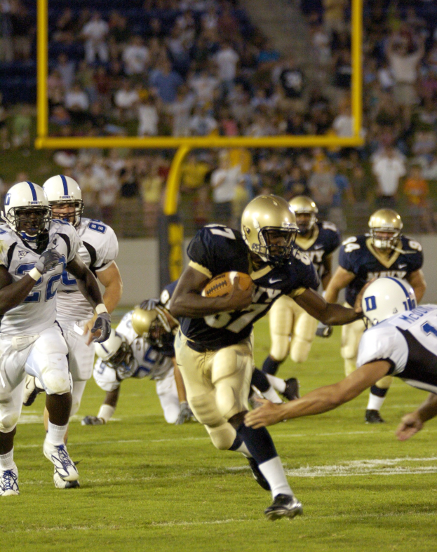 Ein Fußballspieler in Uniform rennt mit dem Ball, während er von Gegnern in Sportkleidung und Helmen verfolgt wird.