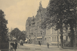 Ein altes Schwarz-Weiß-Foto einer Stadtstraße in Lübeck, Deutschland, das Menschen beim Gehen, einen Pferdewagen, Gebäude mit Fenstern, Bäume, Laternenpfähle und einen Himmel im Hintergrund zeigt, mit dem Text "Stadthallen, Lübeck, Deutschland" unten.