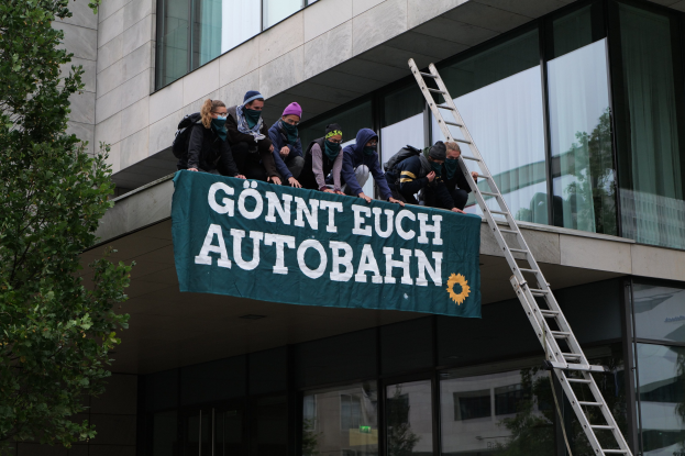 Eine Gruppe von Menschen auf dem Dach eines Gebäudes mit einer Fahne, einem Baum links und Glasfenstern im Hintergrund, wahrscheinlich protestierend für die deutsche Regierung.