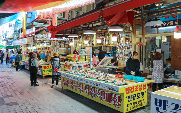 Ein belebter Straßenmarkt in Seoul, Südkorea, mit Menschen, die gehen, Tischen mit verschiedenen Gegenständen und Gebäuden unter einem klaren blauen Himmel.
