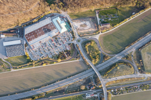 Luftaufnahme eines großen Gebäudes auf einem Feld, umgeben von Straßen, Fahrzeugen, Bäumen und Gras, mit einem Firmenlogo, das darauf hinweist, dass es eine Firmenzentrale ist.