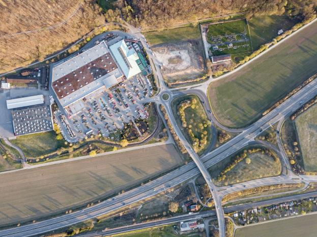 Luftaufnahme eines großen Gebäudes auf einem Feld, umgeben von Straßen, Fahrzeugen, Bäumen und Gras, mit einem Firmenlogo, das darauf hinweist, dass es eine Firmenzentrale ist.