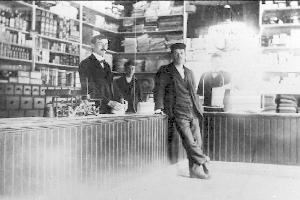 Drei Männer stehen an einem Tresen in einem Geschäft, umgeben von verschiedenen Gegenständen, mit einem Bücherregal und Gegenständen im Hintergrund, die scheinbar einen Raubüberfall durchführen.