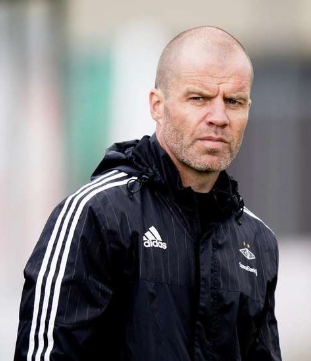 Ein kahlköpfiger Mann in einer blauen Jacke mit weißen Streifen und einem weißen Kragen steht auf einem Fussballfeld, die Hände gefaltet und mit einem ernsten Gesichtsausdruck.