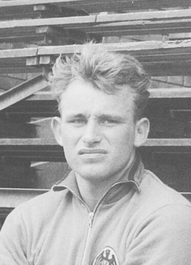 Schwarzes und weißes Foto eines Mannes in einer Jacke vor gestapelten Holzplanken, der legendäre deutsche Fußballspieler.