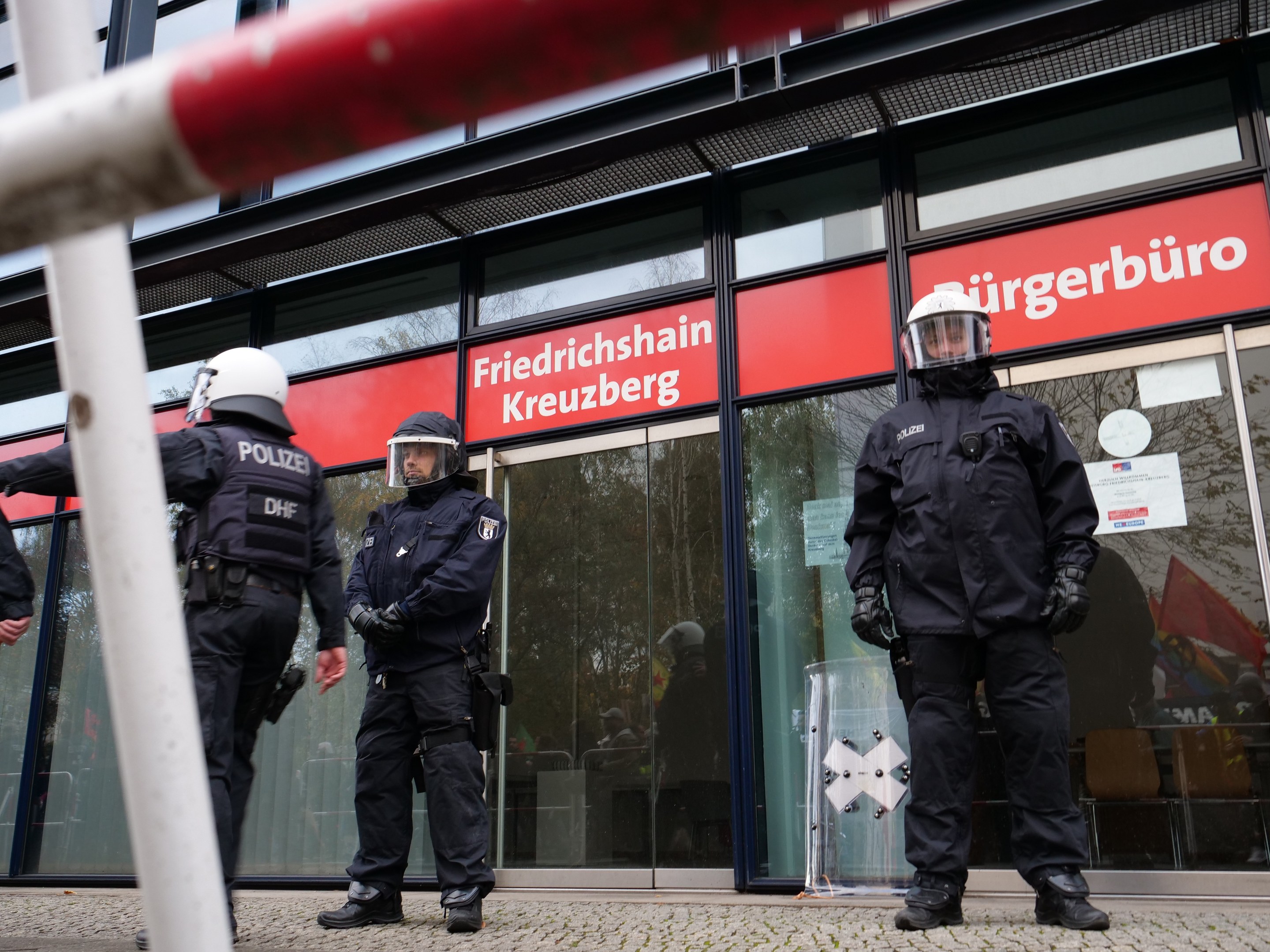 Polizeibeamte in Uniform stehen vor einem Glasgebäude, mit einem Pfahl links und Schildern mit der Aufschrift "Friedrichshain Kreuzberg" im Hintergrund.