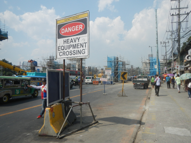 Busy street with a "Danger Heavy Equipment Crossing" sign, vehicles, pedestrians, electric poles, construction buildings, trees, and a cloudy sky.