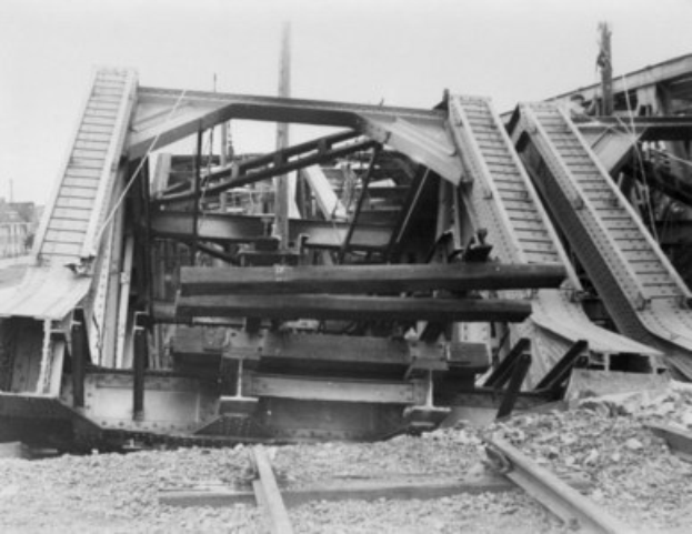 Schwarz-weiß-Foto einer Schienenstrecke, die zu einer zerfallenden Brücke mit gebrochenen Geländern und Schutt führt, umgeben von Gebäuden unter einem klaren Himmel.