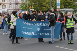 Eine bunte Gruppe von Menschen marschiert eine Straße entlang, hält ein "March for Science Frankfurt am Main"-Schild hoch, mit Bäumen, Pfählen, Schildern, Gebäuden und einem klaren blauen Himmel im Hintergrund.