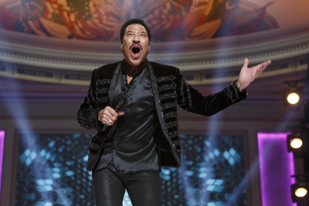 Lionel Richie performs bei den Grammys, trägt ein schwarzes Kleid und hält ein Mikrofon auf einer hell erleuchteten Bühne mit einem Gemälde im Hintergrund.