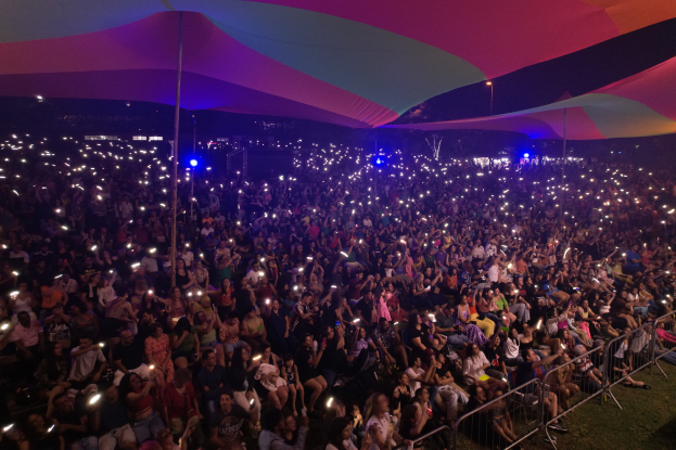 Eine große Menschenmenge sitzt auf dem Rasen vor einem Zelt in der Nacht, beleuchtet von Festival-Lichtern, mit Geländern und Pfählen im Vordergrund und einem bunten Tuch oben.
