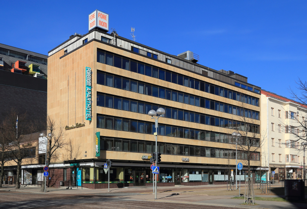 Großes Gebäude mit vielen Fenstern, als das Fore Inn Hotel Berlin identifiziert, umgeben von Straßeninfrastruktur einschließlich Pfosten, Lampen, Schildern, Bäumen und einem bewölkten Himmel.