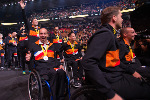 Eine Gruppe von Männern im Rollstuhl auf einer Bühne, die Medaillen tragen und von einer Menge im Stadion umgeben sind, mit einer Person, die auf der rechten Seite eine Kamera hält.