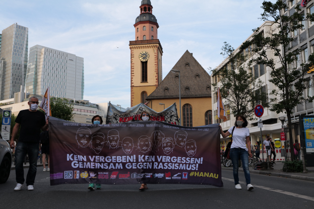 Eine Gruppe von maskierten Menschen geht eine Straße entlang, hält ein Banner hoch, mit einem geparkten Auto auf der linken Seite, Gebäuden und Bäumen im Hintergrund und einem klaren blauen Himmel.
