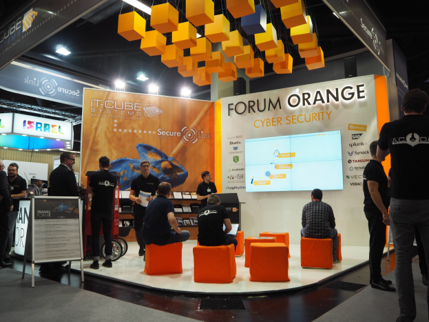 Messe-Stand für IT-Cube Systems Forum Orange Cyber Security mit Menschen, die stehen und sitzen, einer Tafel mit Text, einem Bildschirm, Regalen mit Gegenständen, Schildern, Decke und Lampen.